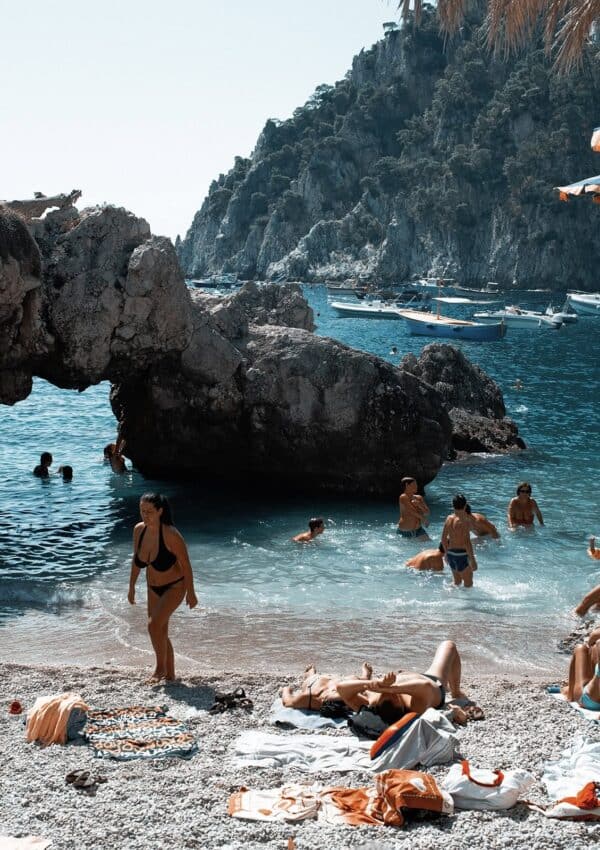 Tourists relaxing on the beach and swimming in the water near an arch rock at Marina Piccola beach in Capri, Italy.