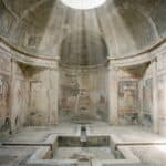 Interior photograph of the Stabian Baths in Pompeii, circular domed ceiling with oculus, dusty shafts of light falling through the opening, pale stone and plaster wall.