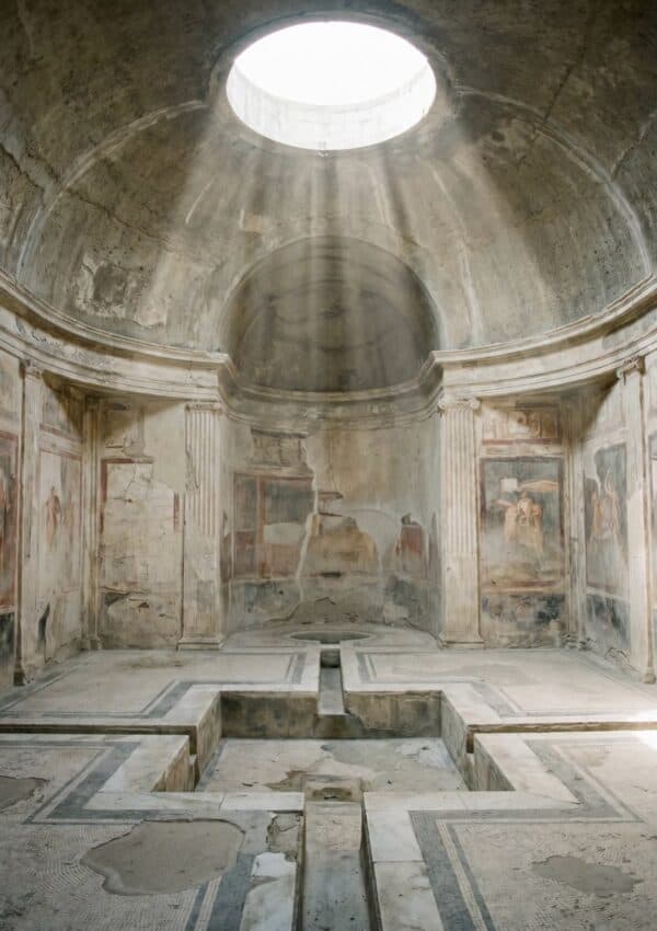 Interior photograph of the Stabian Baths in Pompeii, circular domed ceiling with oculus, dusty shafts of light falling through the opening, pale stone and plaster wall.