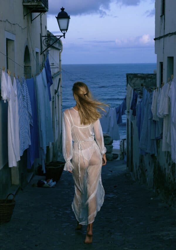 Victoria Van Der Berg walking away down a narrow Capri alley at dusk, white laundry hanging overhead, sea glimpsed at the end of the lane