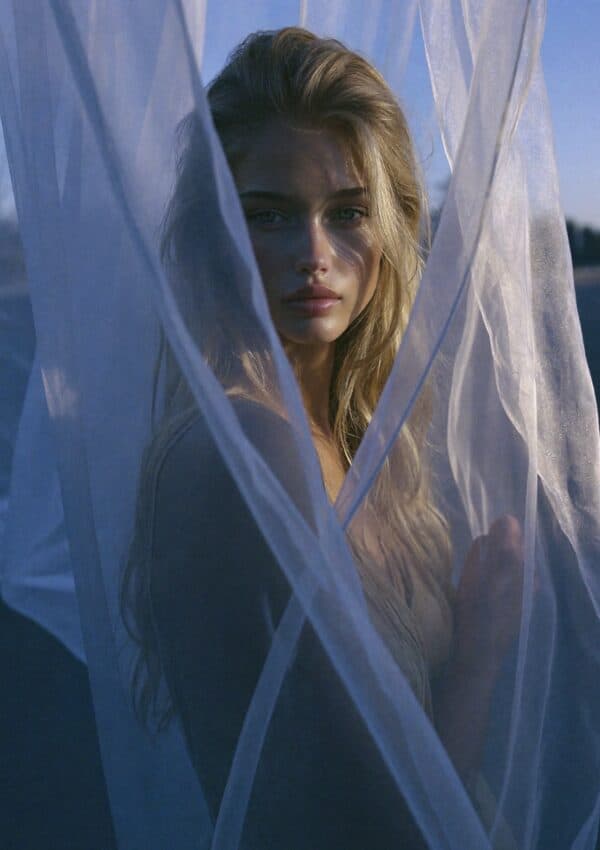 Victoria Van Der Berg's face emerging through white sheer fabric against a vivid blue sky in Capri, making direct eye contact