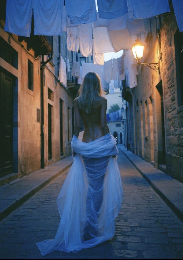 Victoria Van Der Berg walking away down a Capri cobblestone street at night beneath clotheslines of laundry, draped in white sheer fabric