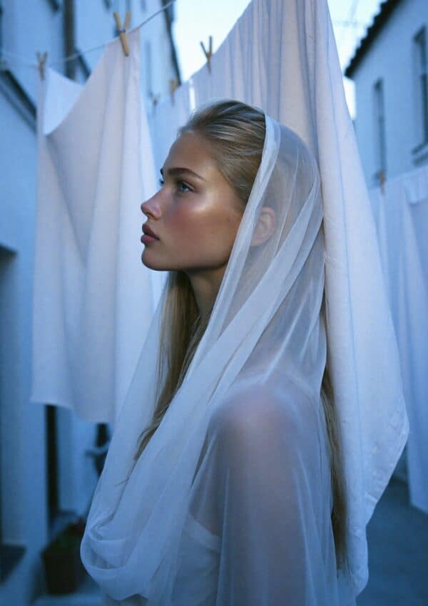 Victoria Van Der Berg in profile, veiled by a hanging white bedsheet in a narrow Capri alley at blue hour, clothespins visible above