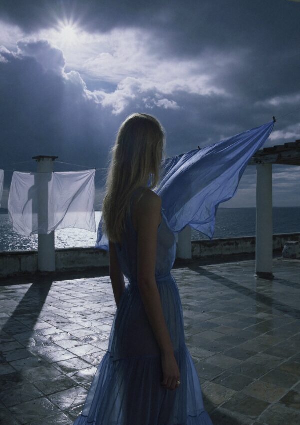 Victoria Van Der Berg on a Capri rooftop terrace in a blue dress, with a stormy sky and sea behind her and blue laundry billowing in the wind