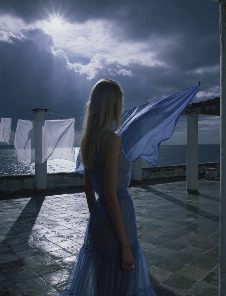 Victoria Van Der Berg on a Capri rooftop terrace in a blue dress, with a stormy sky and sea behind her and blue laundry billowing in the wind