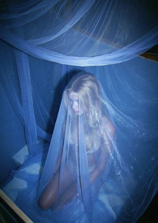 Victoria Van Der Berg sitting inside a blue mosquito net bed canopy in a Capri room, sheer veil draped over her face, bathed in blue light