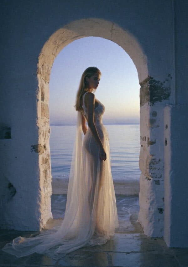 Victoria Van Der Berg standing in an ancient whitewashed stone archway at twilight in Capri, sheer white trailing dress, sea and pink-blue dusk sky visible through the arch