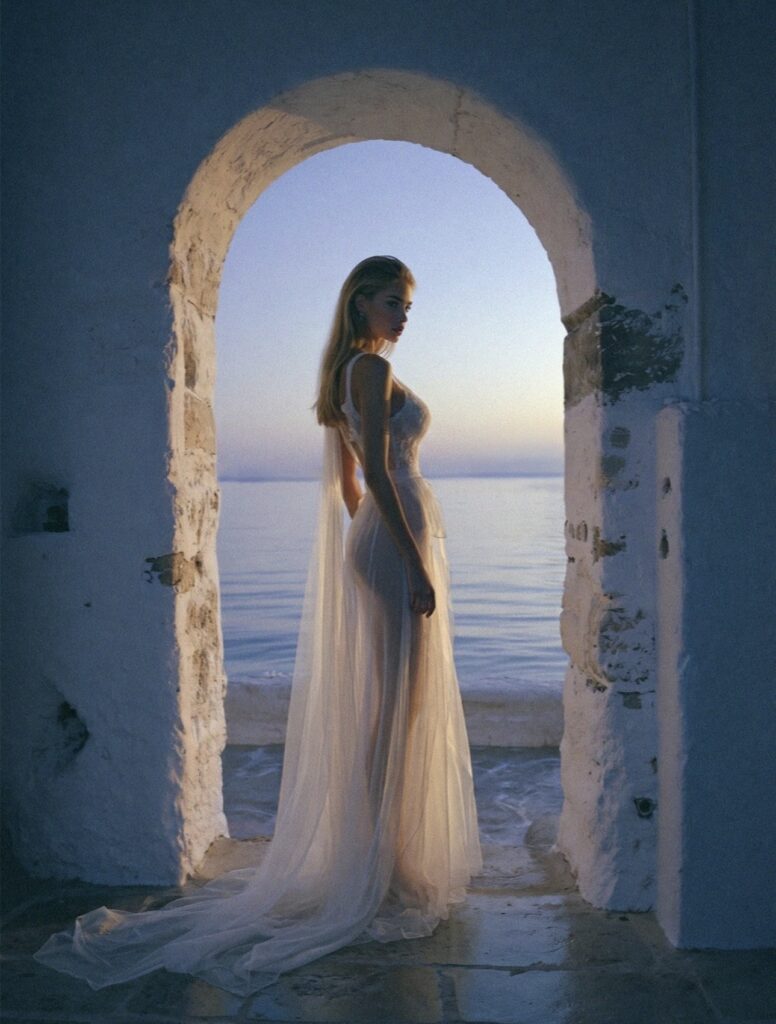 Victoria Van Der Berg standing in an ancient whitewashed stone archway at twilight in Capri, sheer white trailing dress, sea and pink-blue dusk sky visible through the arch