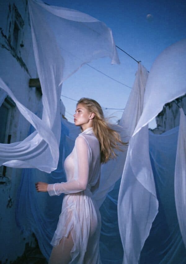 Victoria Van Der Berg surrounded by white sheets billowing in the wind on a Capri rooftop at dusk, sheer white dress, deep blue sky with crescent moon visible