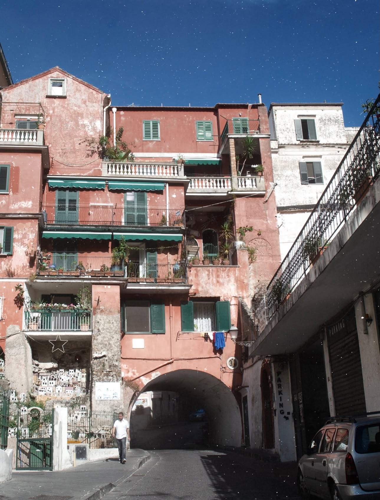 An Amalfi street with a terracotta pink building green shutters and a miniature presepe village display built into the stone wall beside the archway