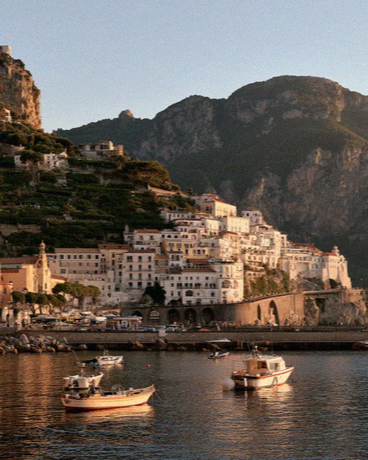 The town of Amalfi seen from the sea at golden hour, houses stacked on the cliff above the harbour