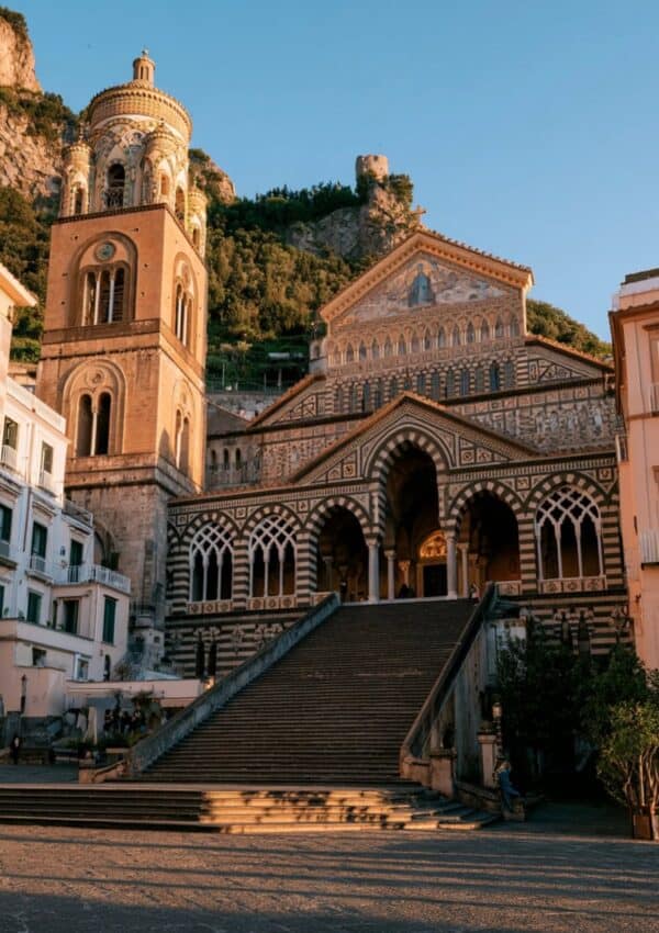 The black and white striped Arab-Norman facade of the Duomo di Amalfi Cathedral of Sant'Andrea rising above the grand stone steps of Piazza del Duomo at golden hour
