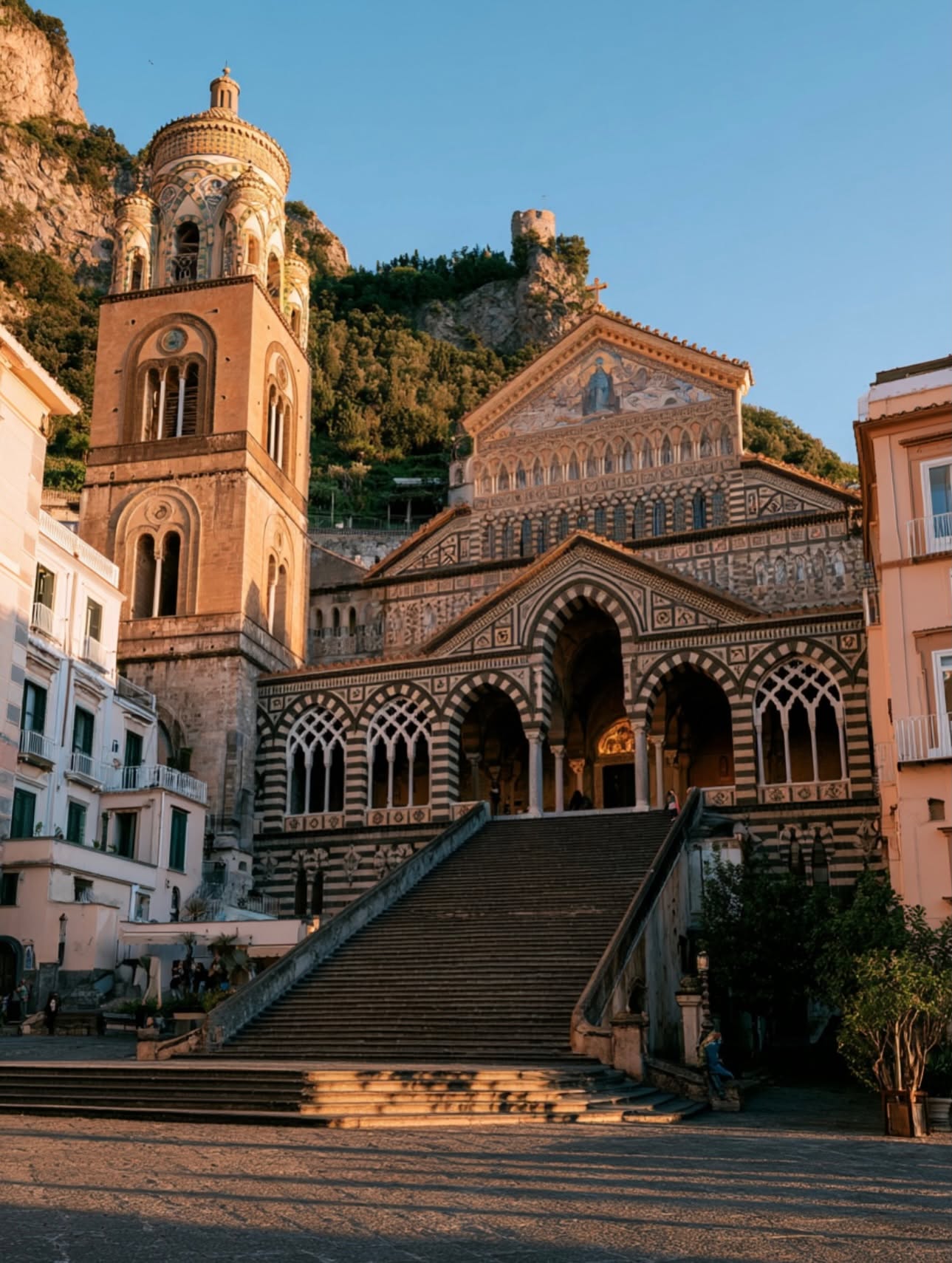 The black and white striped Arab-Norman facade of the Duomo di Amalfi Cathedral of Sant'Andrea rising above the grand stone steps of Piazza del Duomo at golden hour
