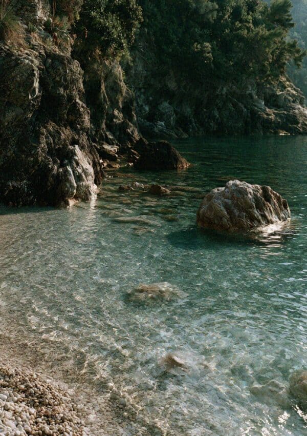 A pebbled cove shore and clear turquoise water beneath wooded cliffs near Portofino