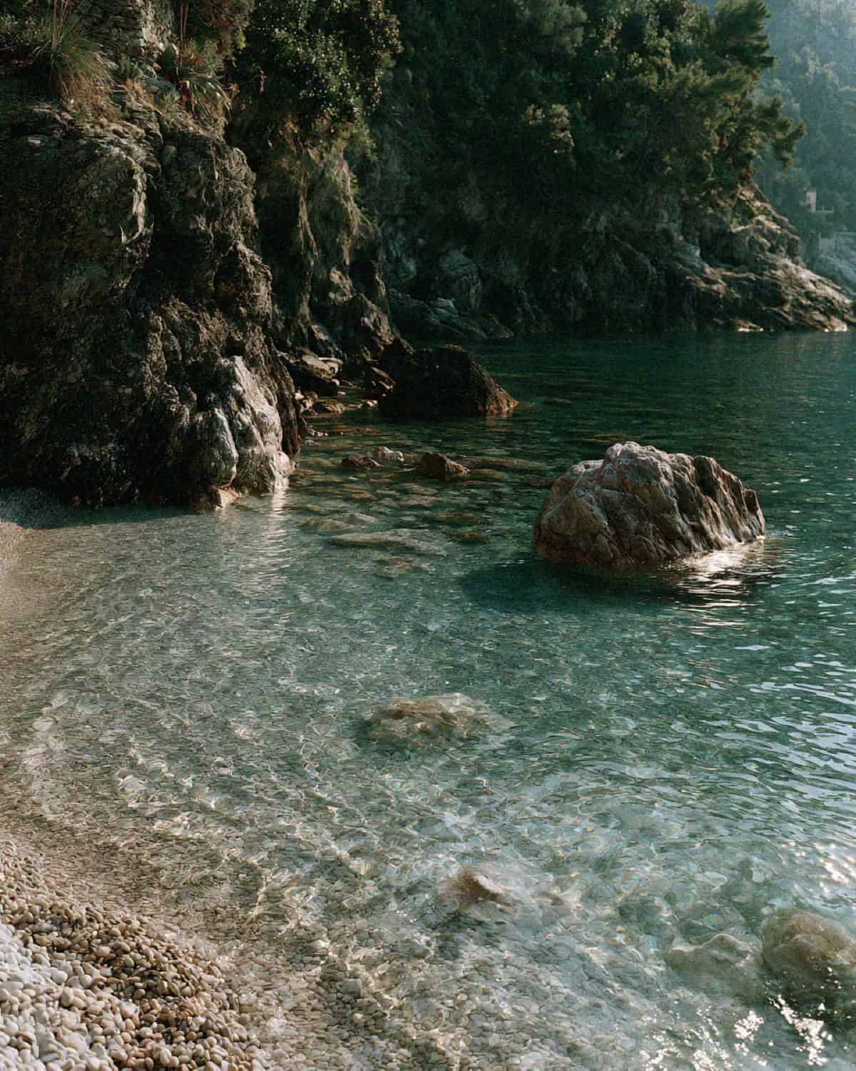 A pebbled cove shore and clear turquoise water beneath wooded cliffs near Portofino