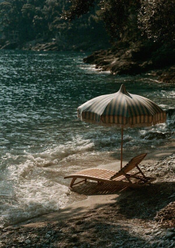 A striped fringed umbrella and wooden sun lounger on the pebble shore of a Portofino cove