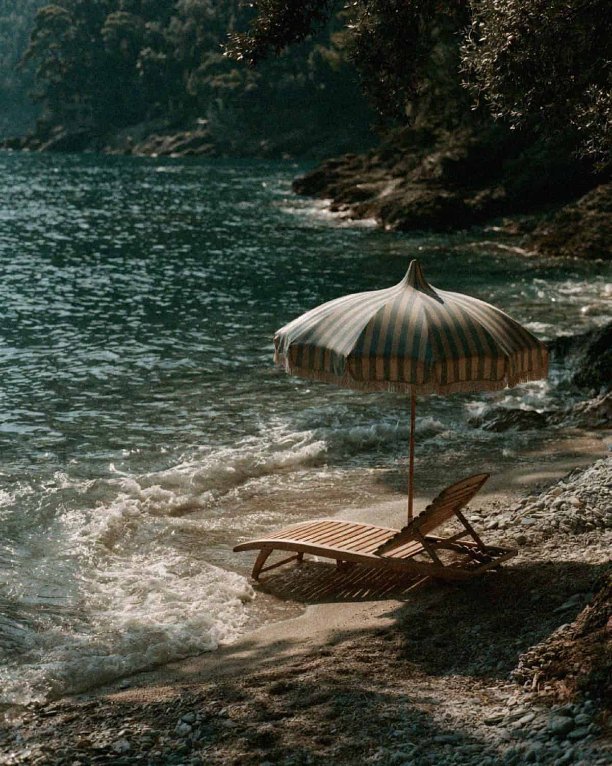 A striped fringed umbrella and wooden sun lounger on the pebble shore of a Portofino cove