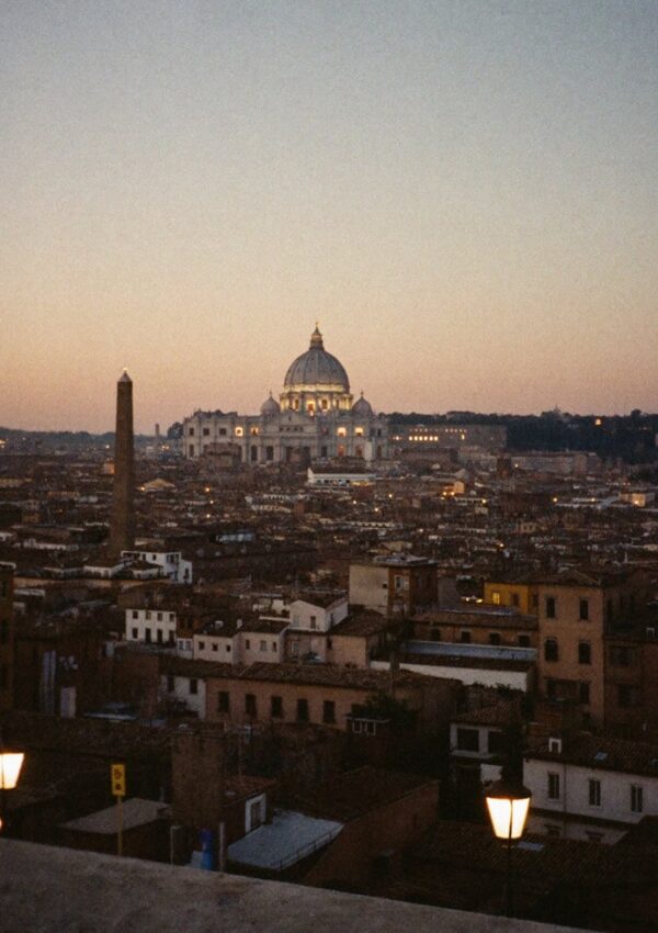 The dome of Saint Peters rising above the rooftops of Rome at dusk seen from a terrace with street lamps