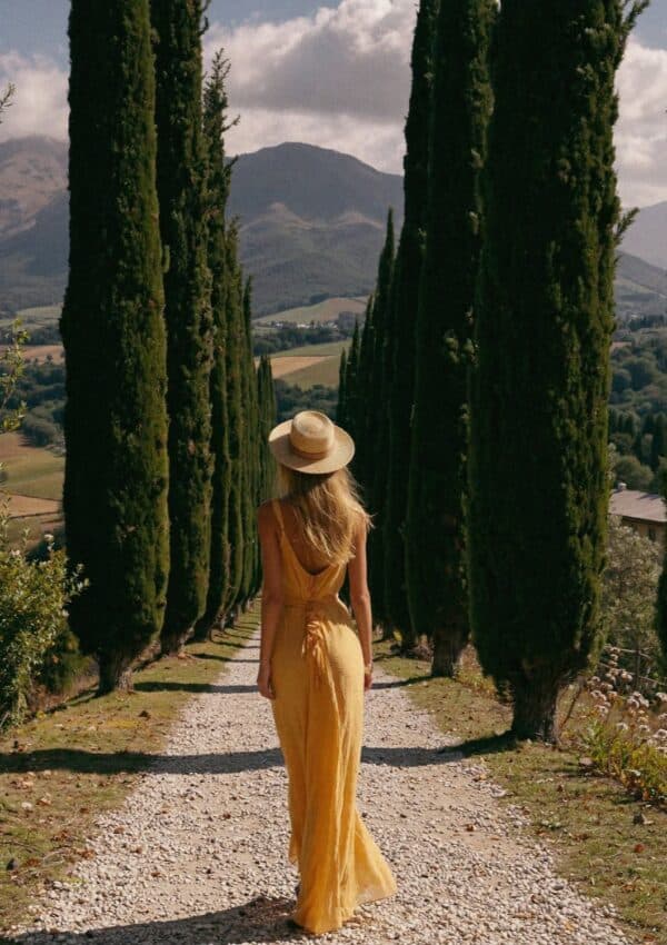 Victoria Van Der Berg walking a cypress-lined gravel path in a yellow dress in Tuscany