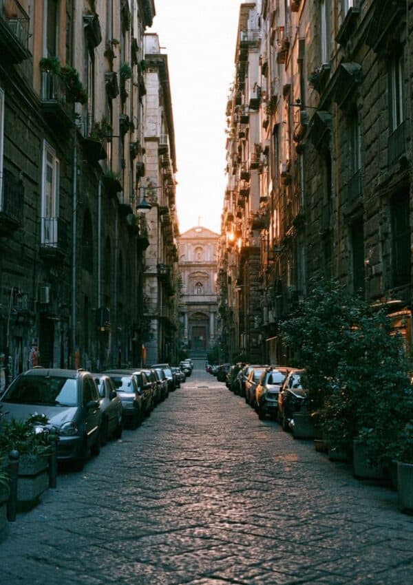 A narrow Naples old town street at sunset with parked cars and a church facade catching the last light