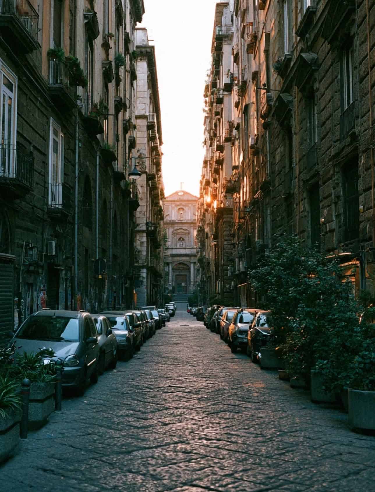 A narrow Naples old town street at sunset with parked cars and a church facade catching the last light