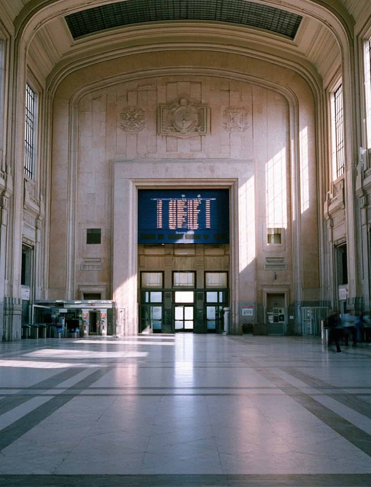 A grand marble station hall with a large departures board, arriving in Naples by train