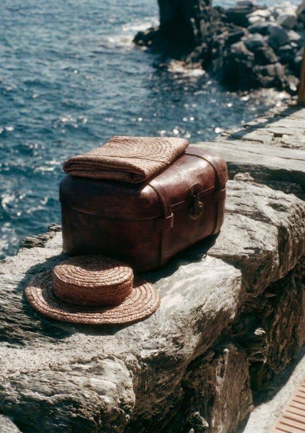 A vintage leather suitcase and a straw hat on the rocks by the sea in Cinque Terre