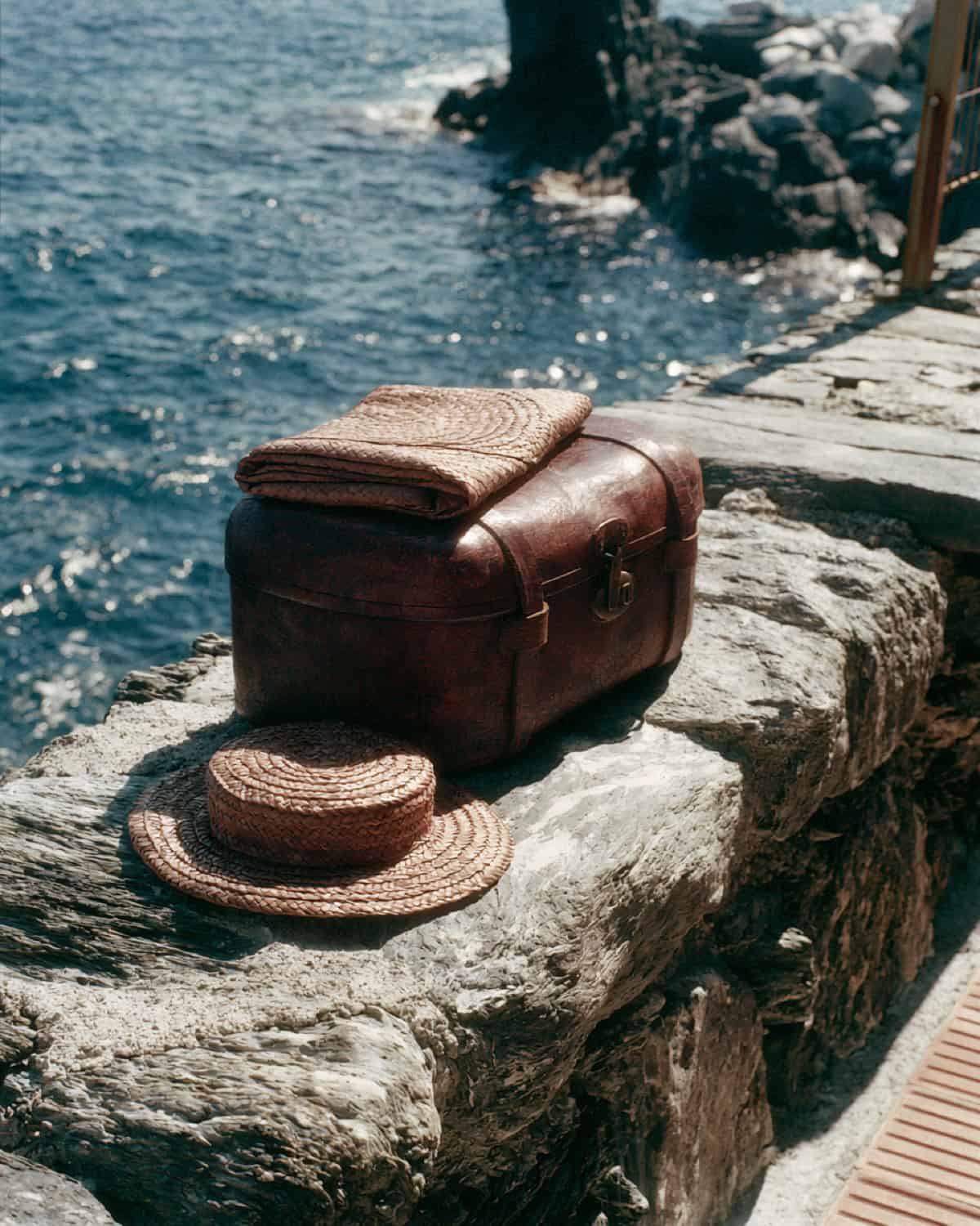 A vintage leather suitcase and a straw hat on the rocks by the sea in Cinque Terre
