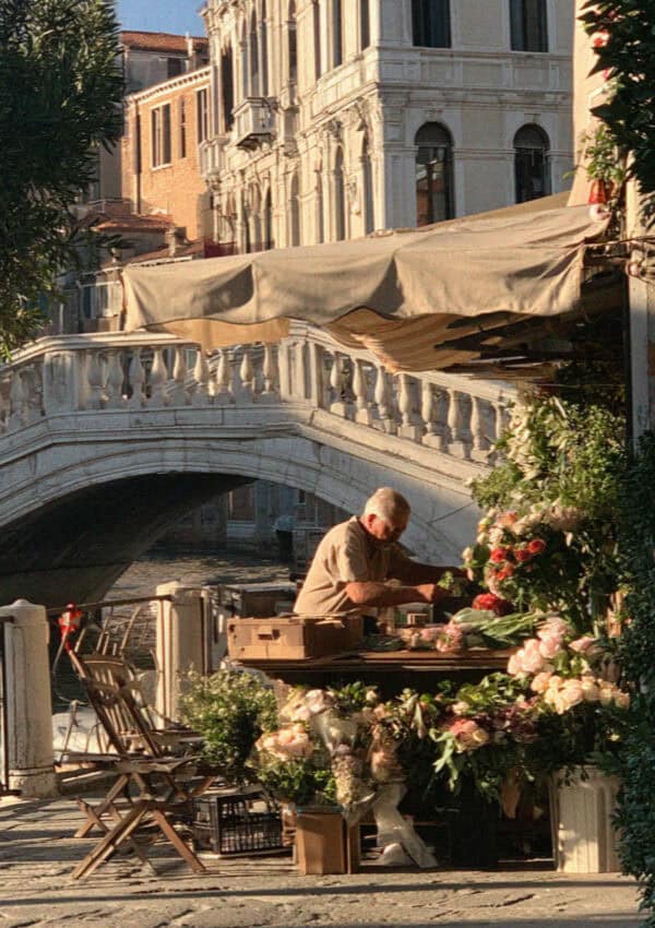 Man arranging flowers at a stall beside a stone bridge on a Venice canal
