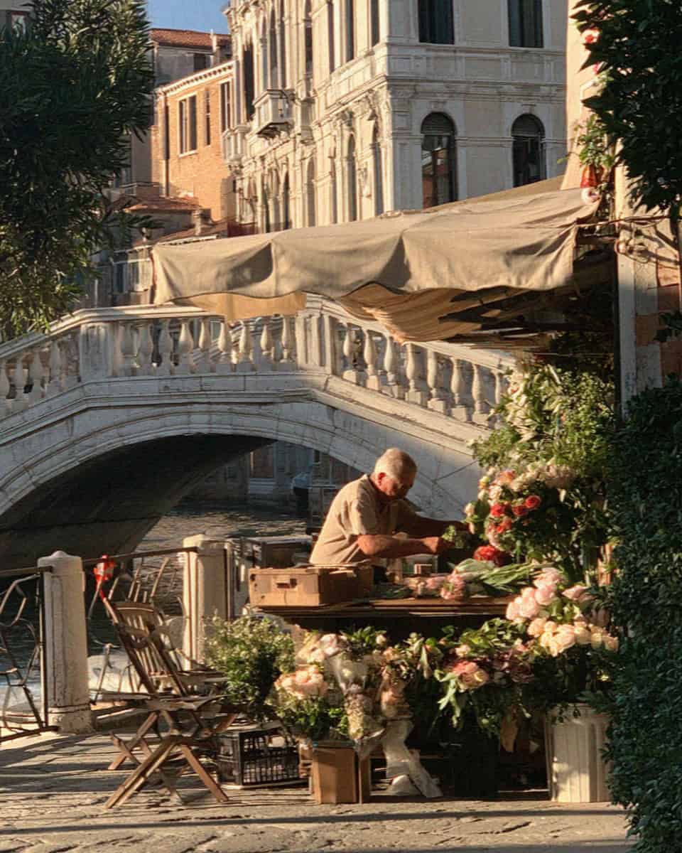 Man arranging flowers at a stall beside a stone bridge on a Venice canal