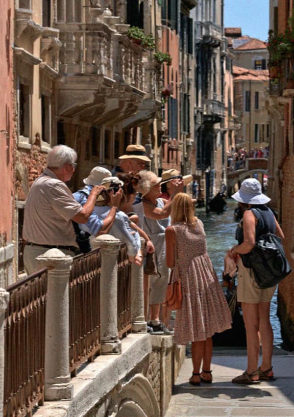 Tourists leaning over a bridge railing to photograph a Venice canal