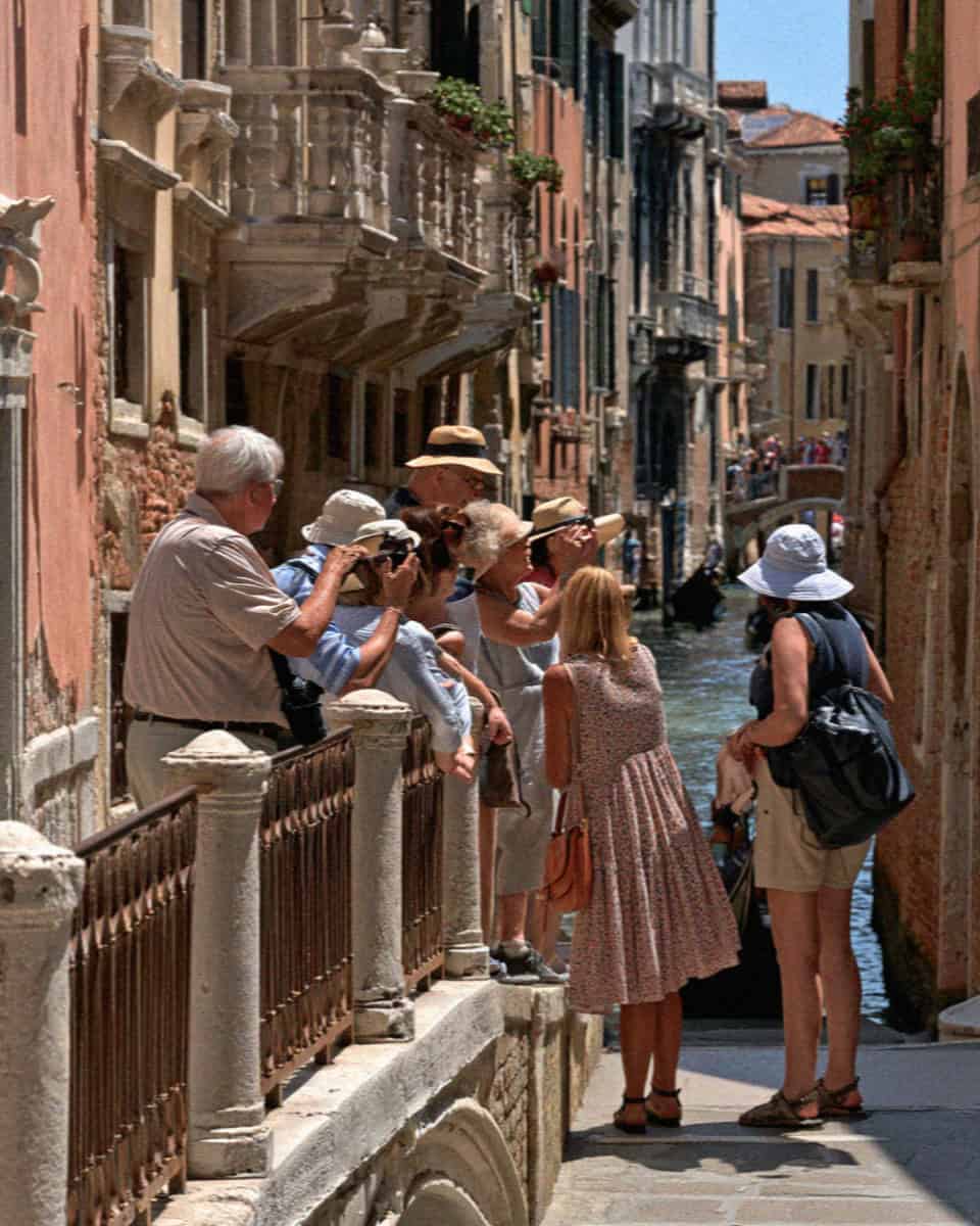 Tourists leaning over a bridge railing to photograph a Venice canal