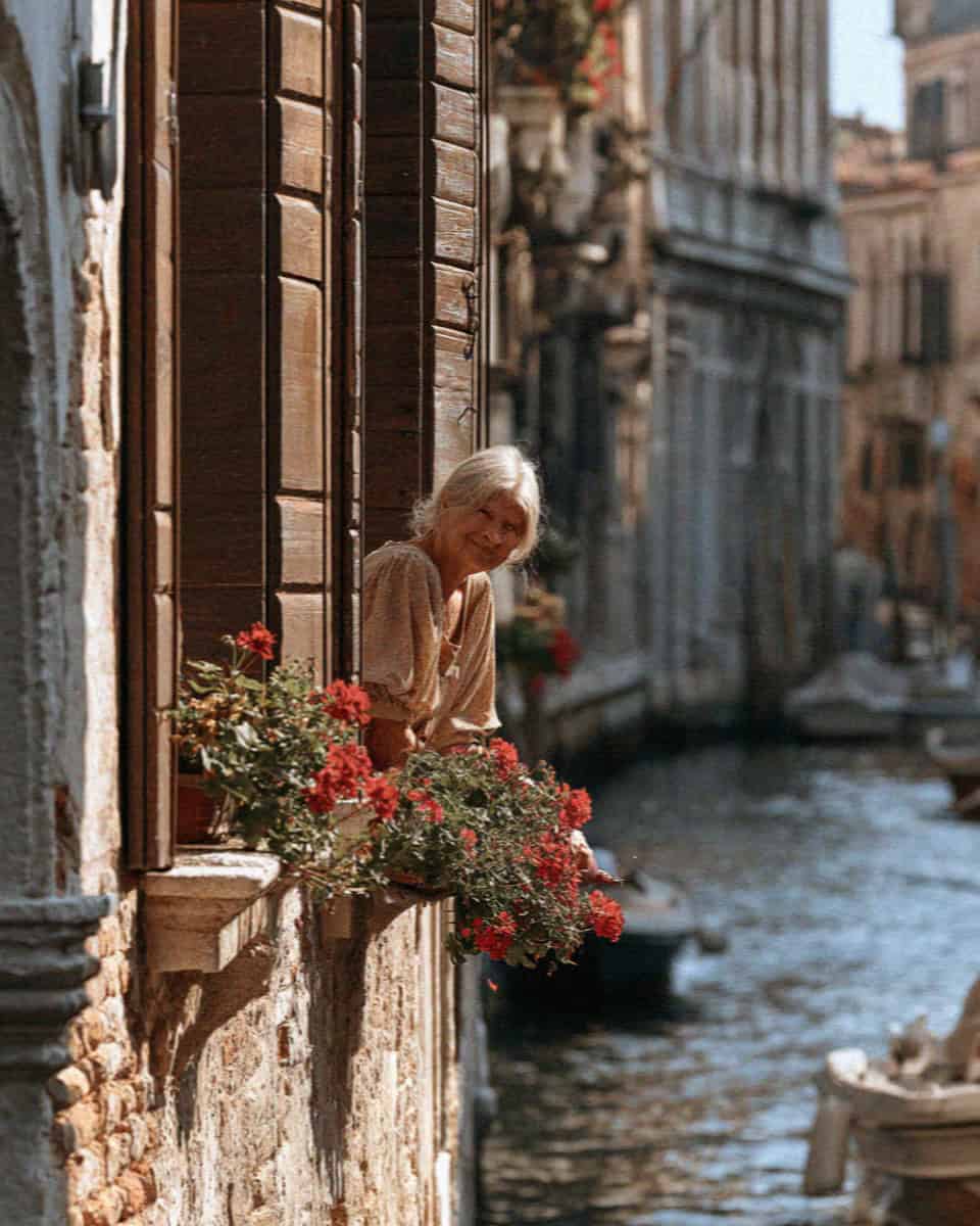Elderly Venetian woman at a window framed by red geraniums above a canal