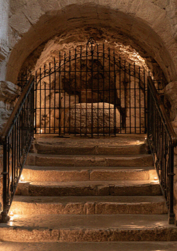 Worn stone steps leading down to a lit crypt behind a wrought-iron grille in Assisi