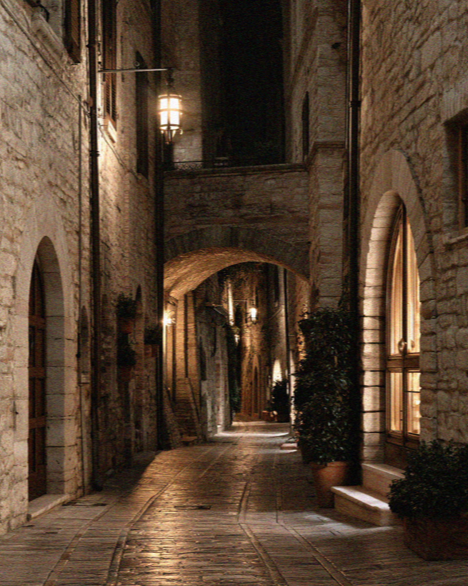 A medieval Assisi lane at night with warm lantern light on pale stone