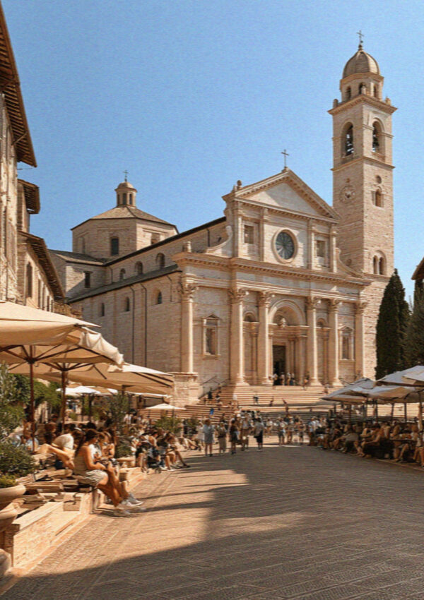 A wide stone square in front of a church in Assisi filling with tour groups and sun umbrellas in hard August light