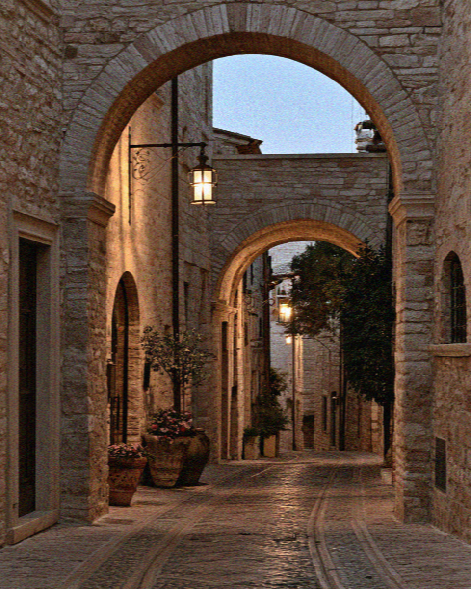 A medieval Assisi alley at twilight with wrought-iron lamps glowing on pale stone