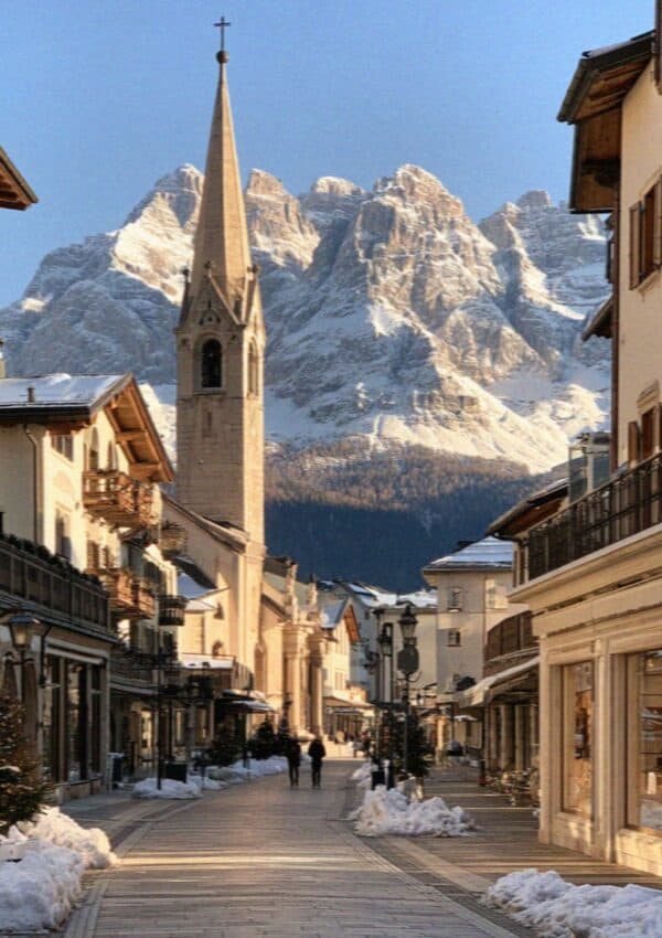 A Dolomites town street with a church spire and snowy peaks in winter