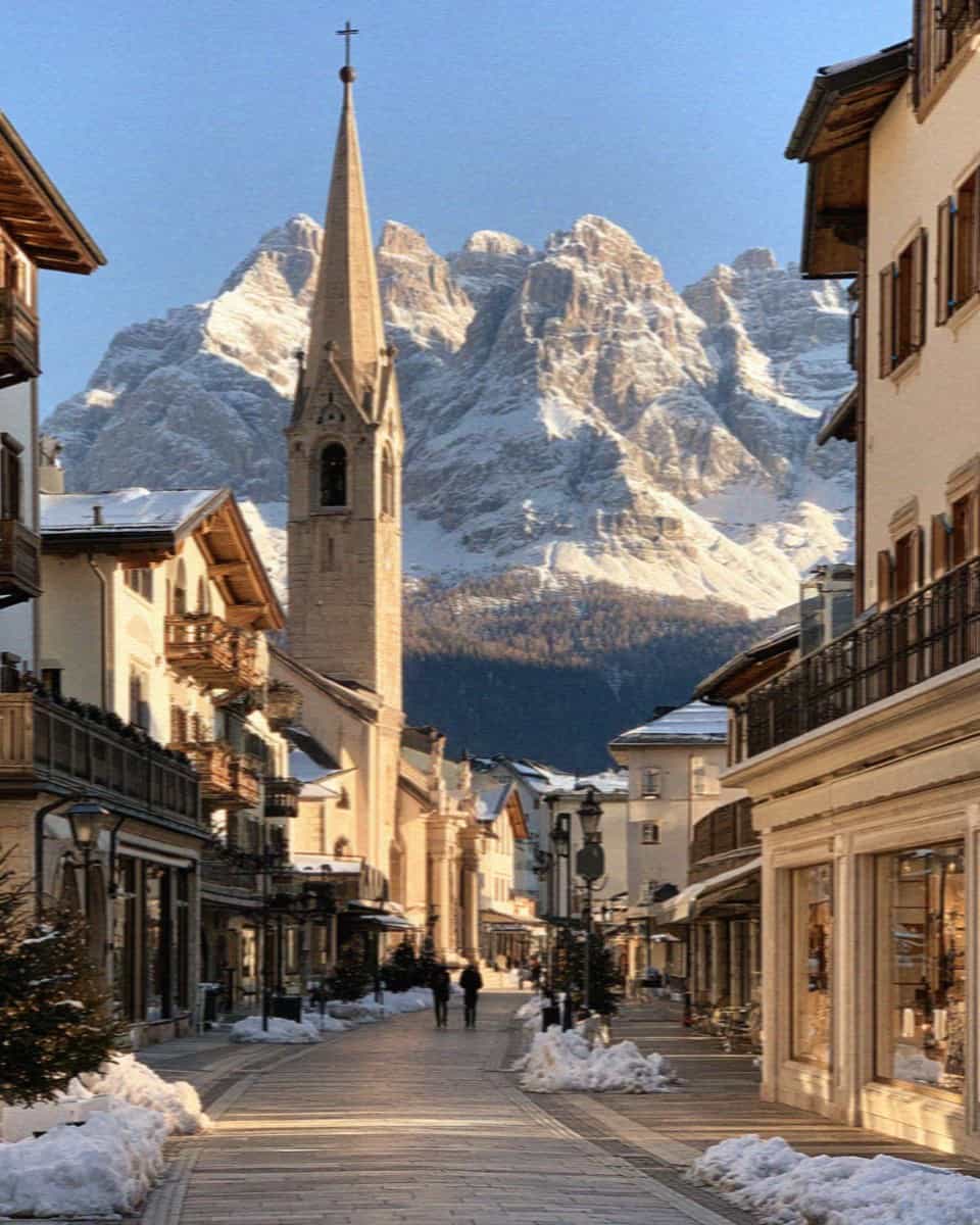 A Dolomites town street with a church spire and snowy peaks in winter