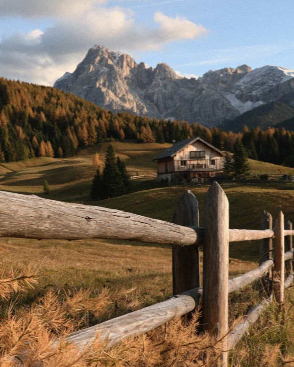 An autumn meadow with a chalet and larches below Dolomite peaks