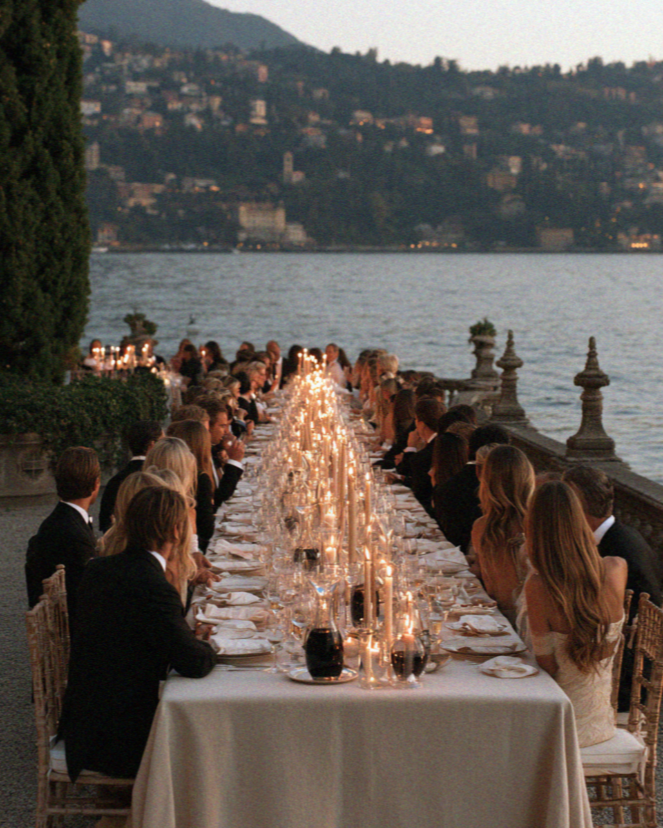 A long communal welcome dinner table set beside the water on Lake Como