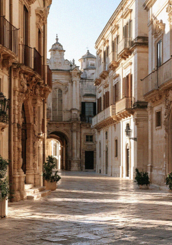 A paved baroque street in Lecce at golden hour, carved palazzo facades on both sides and an archway at the far end