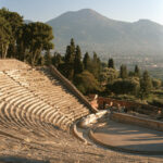 The stone tiers and oval arena of the ancient amphitheatre in Pompeii seen from high on the seating in warm late light, no people