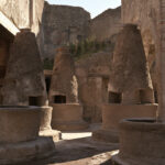 The tall conical volcanic stone grinding mills of an ancient bakery in Pompeii under hard high sun, no people