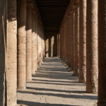 A long colonnaded portico of weathered brick columns in Pompeii throwing repeating shadows across the paving, no people