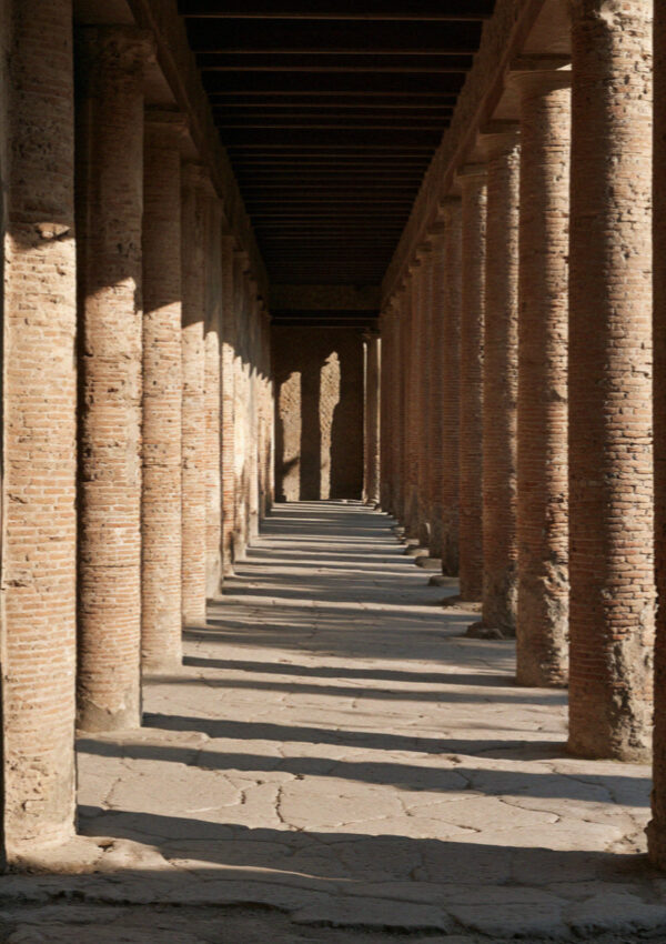 A long colonnaded portico of weathered brick columns in Pompeii throwing repeating shadows across the paving, no people