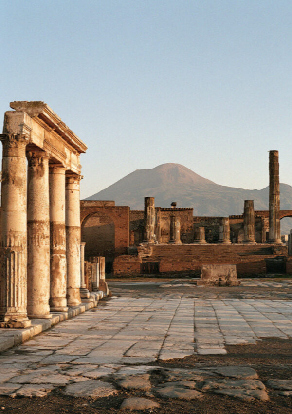 A vast empty ancient forum in Pompeii at opening time, broken columns and bleached paving with Vesuvius pale on the horizon, no people