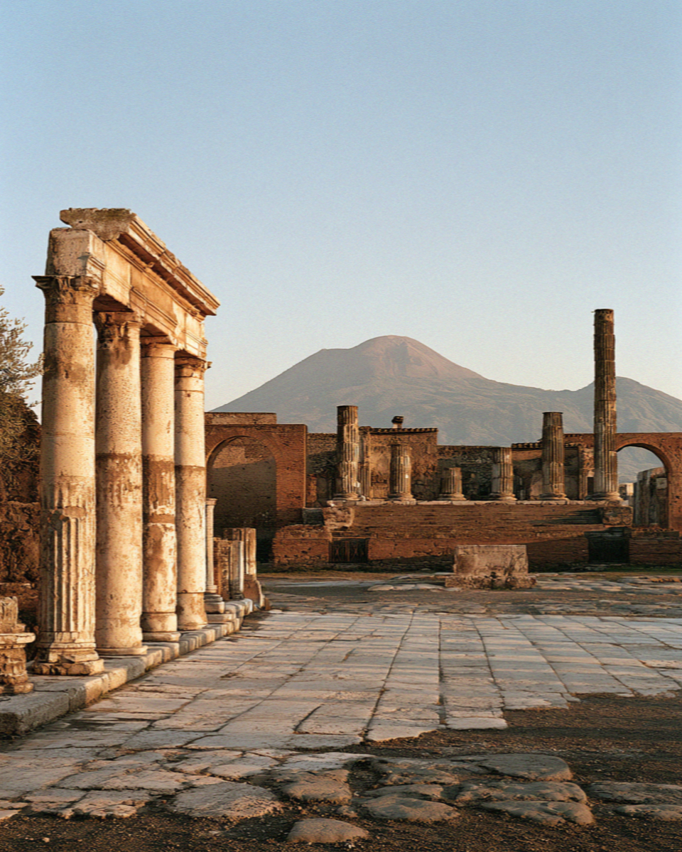 A vast empty ancient forum in Pompeii at opening time, broken columns and bleached paving with Vesuvius pale on the horizon, no people