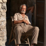 An older weathered site custodian sitting in a shaded doorway in Pompeii with keys and a folding chair