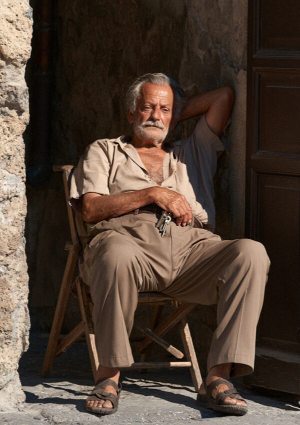 An older weathered site custodian sitting in a shaded doorway in Pompeii with keys and a folding chair